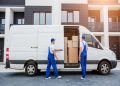 Two movers in blue overalls load cardboard boxes into a white moving van in front of a modern apartment building.