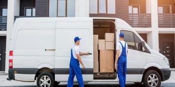 Two movers in blue overalls load cardboard boxes into a white moving van in front of a modern apartment building.