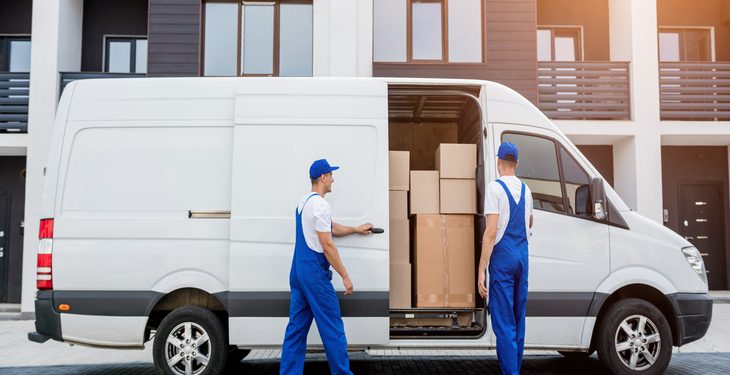 Two movers in blue overalls load cardboard boxes into a white moving van in front of a modern apartment building.