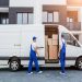 Two movers in blue overalls load cardboard boxes into a white moving van in front of a modern apartment building.